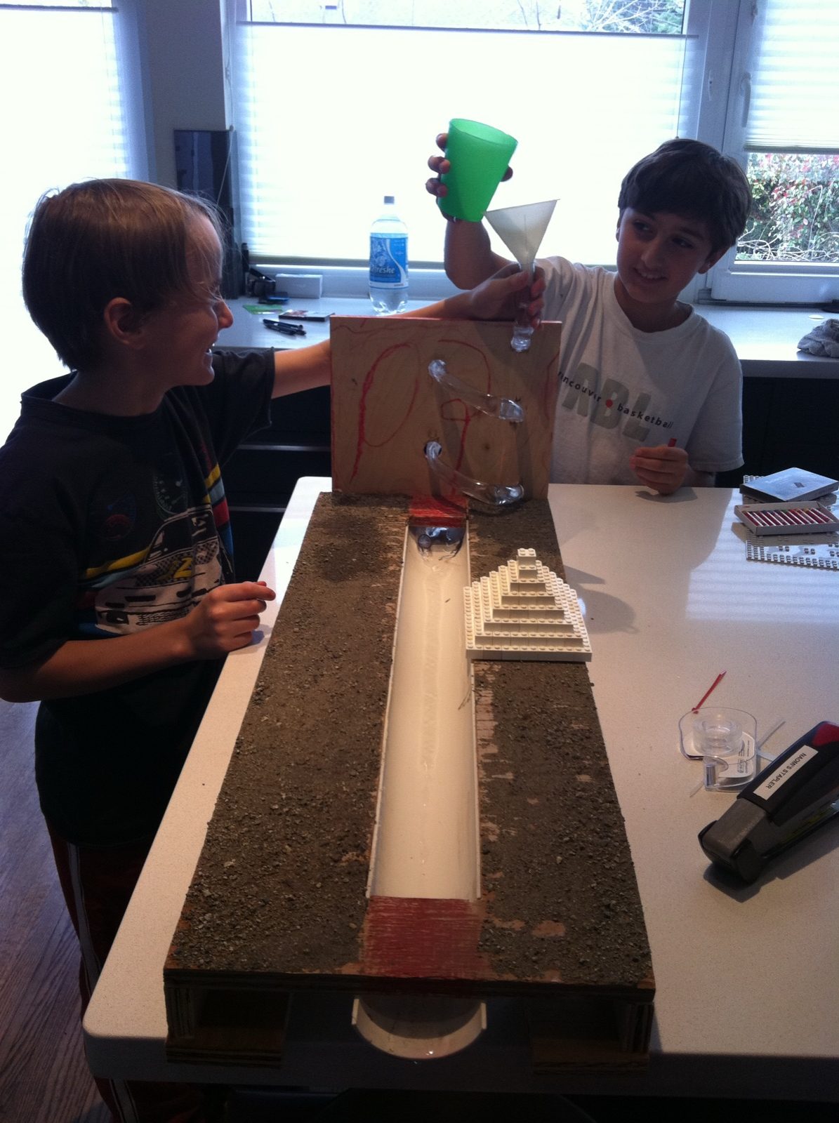Jack and Solly as kids at a kitchen island, pouring water through a funnel into a homemade wooden water-channel model, April 2011