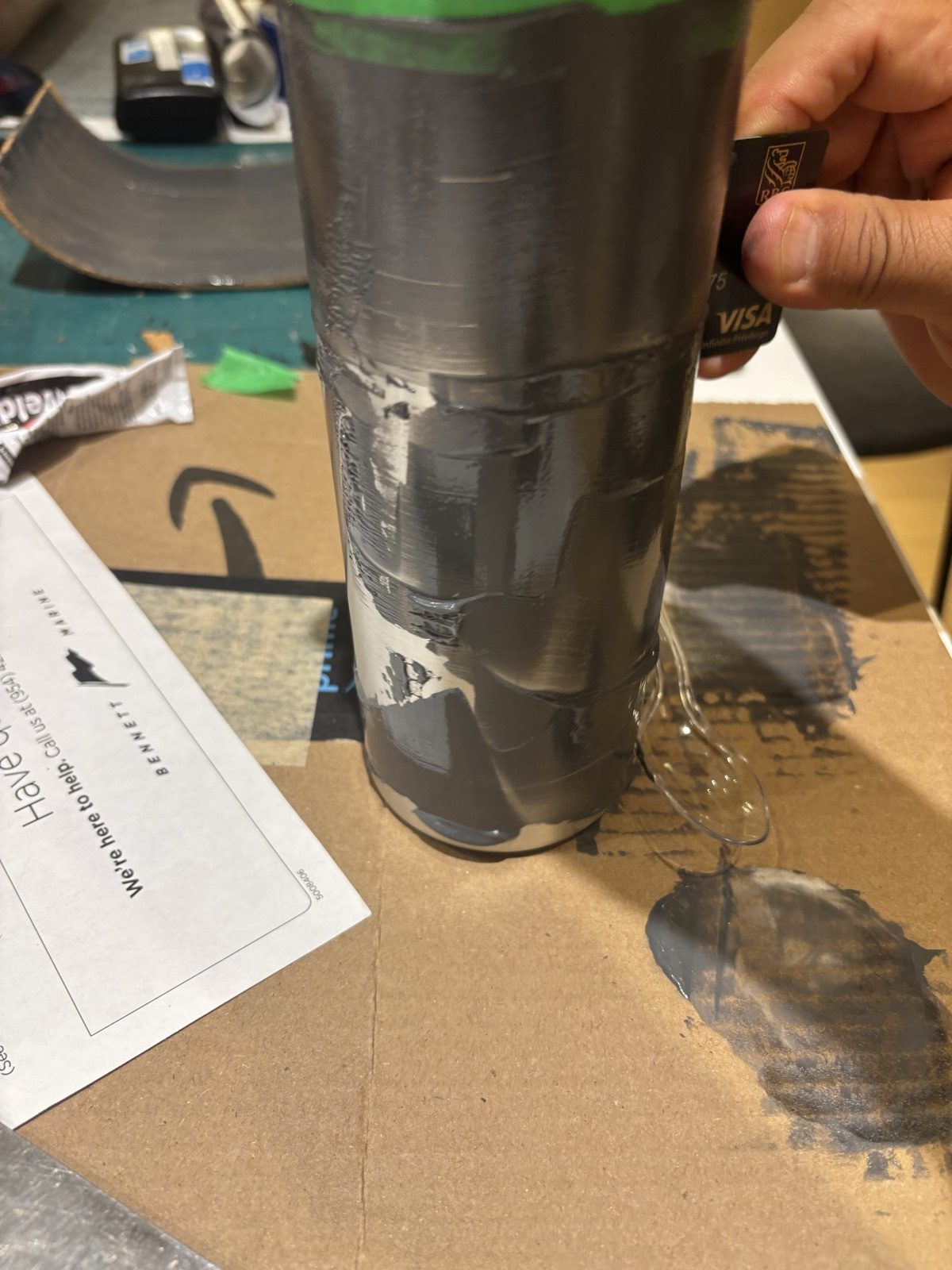 A hand spreading grey epoxy over a steel bottle with the edge of a credit card, on cardboard on a workbench